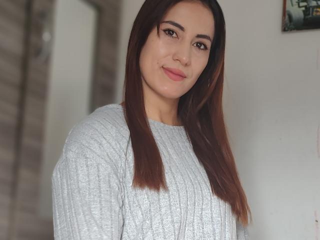SofiaAnal with brown hair, gray knit sweater, and pink lipstick, smiling at the camera, showing her tits slightly, in a well-lit room.