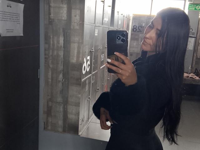 SofiaAnal with long black hair, taking a mirror selfie in a locker room, wearing a black fur coat, holding a black phone, showing her ass in the reflection.