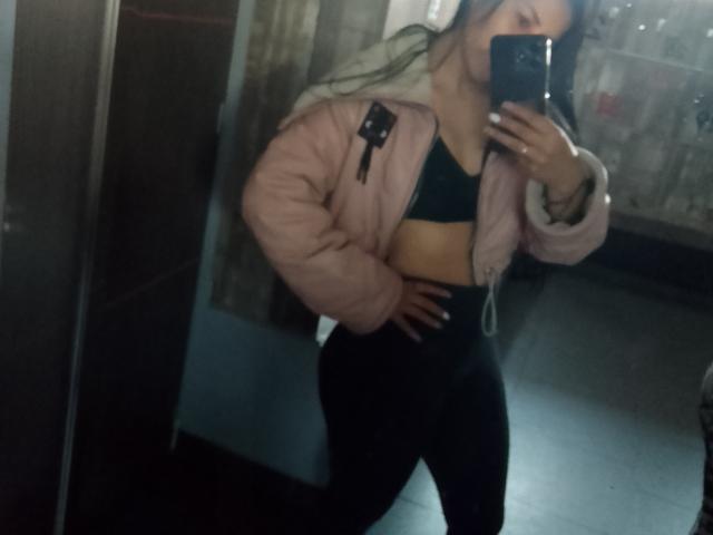 SofiaAnal met lang donker haar, een roze, opvallende jas, een zwarte sportbeha en zwarte leggings met hoge taille, die een spiegelselfie maakt, waarbij haar achterkant en haar hand op haar heup te zien zijn..