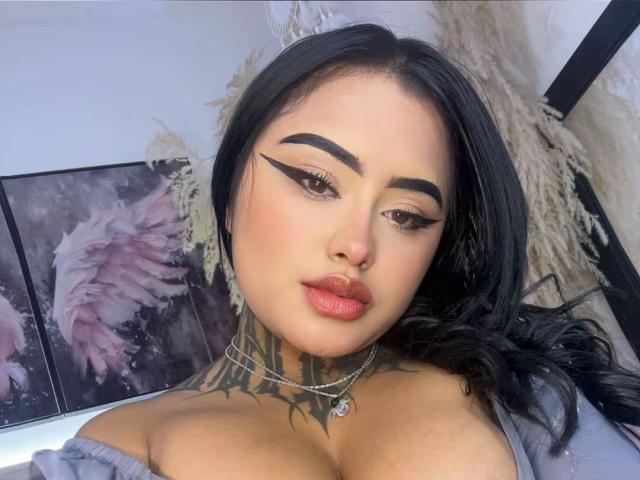 LairaHearvy has black hair, bold winged eyeliner, red lipstick, and a tattoo on her chest. She's wearing a necklace and has large, exposed tits. Her skin is light.