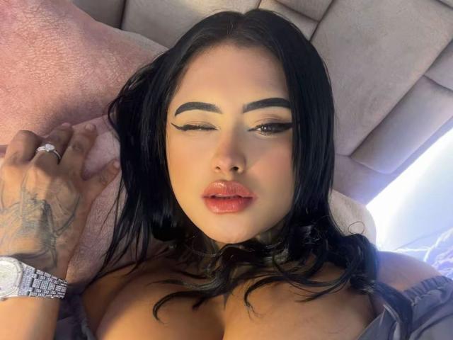 LairaHearvy with winking eye, black hair, bold eyebrows, red lips, cleavage, tattooed hand, ring, and watch, lying on pink pillow.