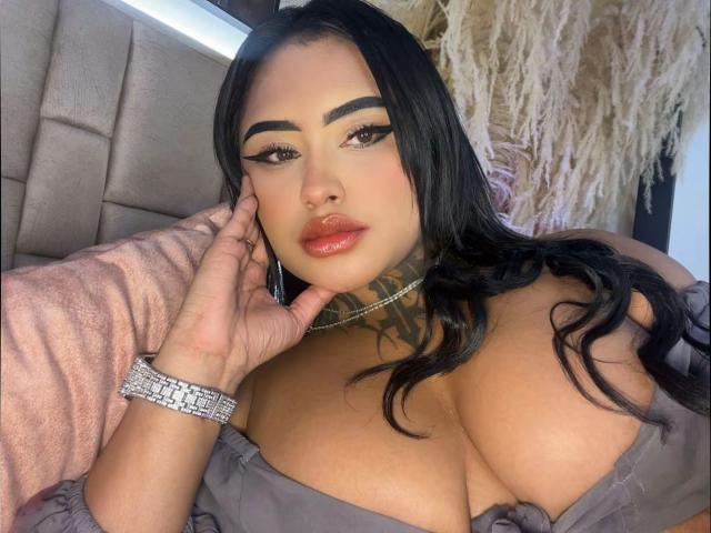 LairaHearvy with black hair, thick eyebrows, and red lips, wearing a grey off-shoulder top, showing her big tits, hand on her cheek, and a diamond bracelet.