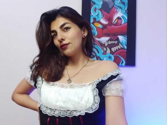 SoyVeronica with wavy dark hair, red lipstick, wearing a black and white lace-off shoulder dress, necklace, standing in front of a colorful painting, smiling slightly.