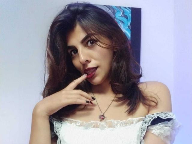 SoyVeronica with medium brown hair, dark eyes, and red lipstick, wearing a white lace off-shoulder top, biting her finger with black nails, and a pink pendant necklace.
