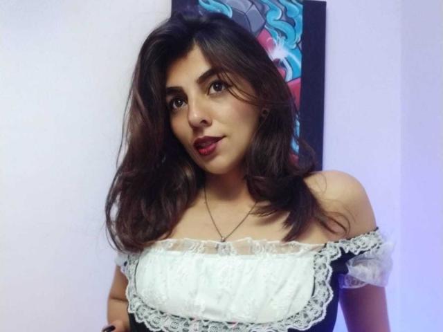SoyVeronica with dark brown wavy hair, wearing an off-shoulder white lace dress, brown eyes, and red lipstick, standing against a white wall with a colorful painting.