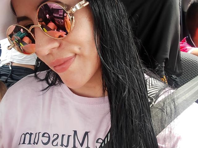 SofiaAnal with wet black hair, wearing reflective gold aviator sunglasses and a pink shirt, smiling slightly. Her pink lips are subtle and her skin is light.