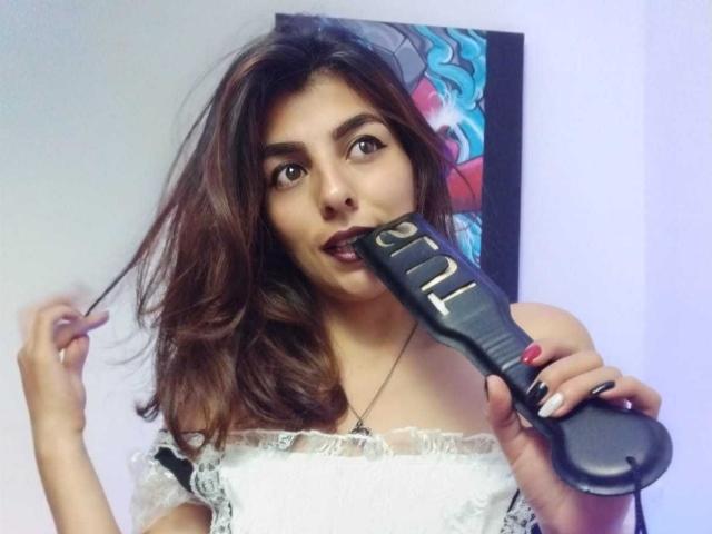 SoyVeronica with brown wavy hair, dark lipstick, holding a black electric shaver to her mouth, wearing a white lace off-shoulder top, fingers with red and white nail polish.