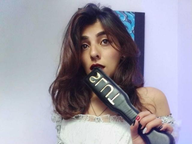 SoyVeronica with wavy brown hair, wearing a white off-shoulder dress, holding a black hairbrush in her mouth, looking at the camera with dark lipstick.