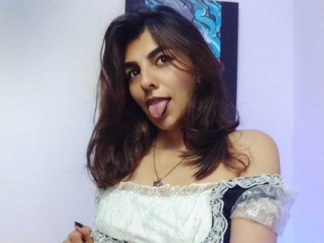 SoyVeronica with medium brown hair, dark makeup, tongue out, wearing an off-shoulder black and white lace dress, necklace, looking playful, holding dress slightly.