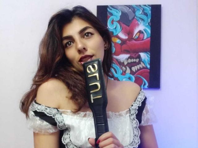 SoyVeronica with long brown hair, wearing a black and white lace maid outfit, holding a black paddle with "PADDLE" in gold letters, and a red, blue, and yellow face painting.