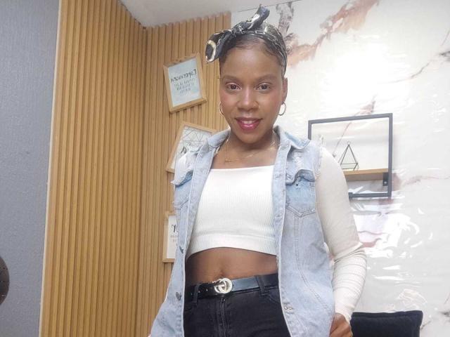 SarahiPusey with dark skin, wearing a denim vest over a white crop top, black high-waisted pants, and a headband with white patterns, standing and smiling.