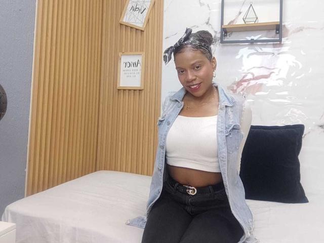 SarahiPusey with dark skin, small breasts, wearing a denim vest, white crop top, and black jeans, smiling, sitting on a bed with a black pillow.
