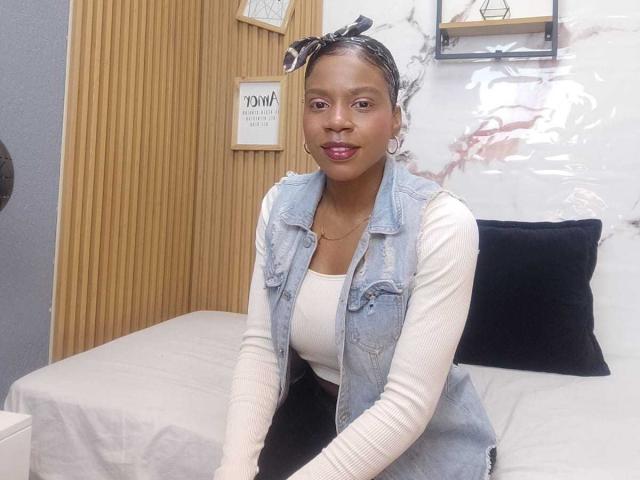 SarahiPusey with dark skin, wearing a denim vest over a white top, black pants, and a black headband, sitting on a bed, smiling with pink lipstick.