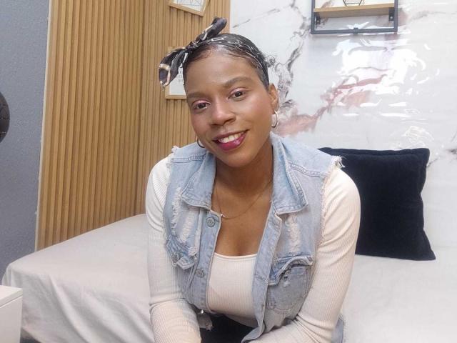 SarahiPusey with dark skin, curly hair in a black headscarf, wearing a denim vest over a white ribbed shirt, smiling with pink lipstick, sitting on a bed.