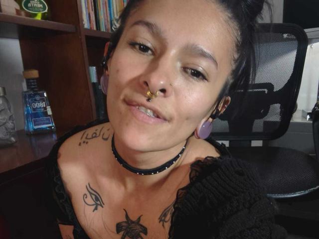 AmelieBlake has tattoos on her chest, nose ring, and purple ear gauges. She's wearing a black choker and looking at the camera with a slight smile.