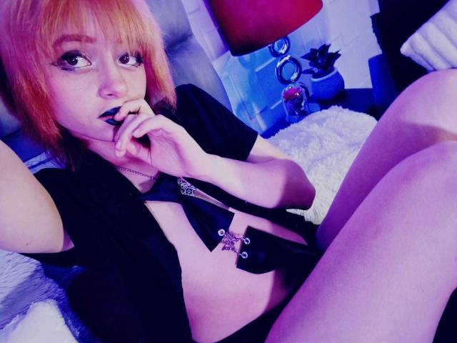 ViolettClay with orange hair, black eyeliner, and lipstick, biting her finger, wearing an open black shirt, exposing her chest, and a necklace, sitting with her legs up.
