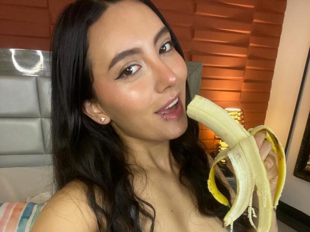 CarlaFranco mit langem, schwarzem Haar, die eine geschälte Banane zu ihrem leicht geöffneten Mund hält und einen verspielten Ausdruck zeigt, wobei ihre kleinen Brüste kaum sichtbar sind. Schlafzimmer, warmes Licht..