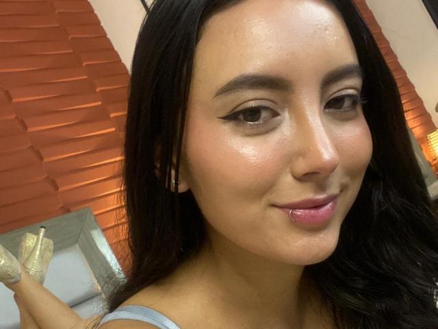 CarlaFranco heeft een glanzende huid, zwart haar, eyeliner met een vleugje, en een roze lipring, draagt een blauwe top, waarbij haar billen iets omhoog zijn, en ze draagt glinsterende hakken..