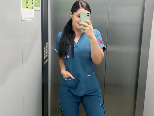 VickyMegan with long black hair, wearing blue nurse scrubs, standing in an elevator, holding a phone in front of her face, hand in pocket, smiling slightly.