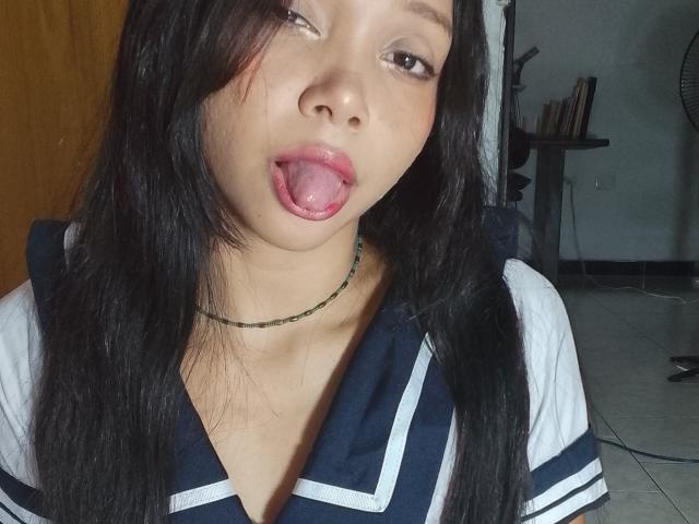 NashaLoule, con lunghi capelli neri, occhi marroni e la lingua rosa in fuori, indossa un'uniforme scolastica blu scuro e bianca e una collana verde, e guarda verso la fotocamera..