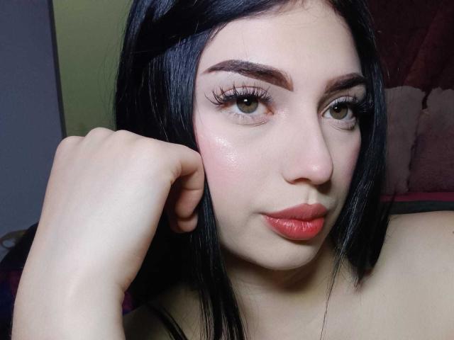 Isabella Rossi a la peau pâle, les cheveux noirs, des sourcils épais, des yeux verts avec de longs cils, et des lèvres rouges. Elle repose son menton sur sa main avec un léger sourire..