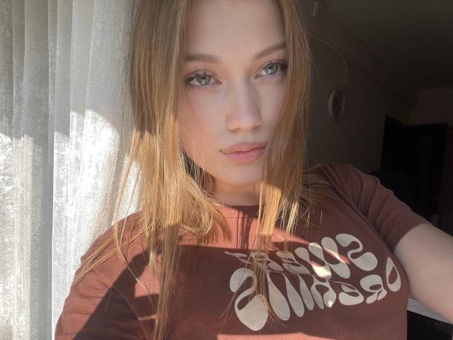 BobbyeWelson with light brown hair, blue eyes, and a cute face, wearing a brown t-shirt, standing in sunlight, showing soft pink lips and a slightly parted mouth.