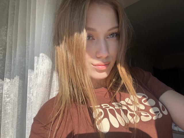 BobbyeWelson with light brown hair, green eyes, and a cute smile, wearing a brown t-shirt with white graphic, sunlit, showing soft light on face and hair.
