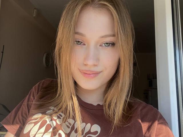 BobbyeWelson with light brown hair, green eyes, and a slight smile, wearing a brown T-shirt, sunlight in the background, looking directly at the camera.