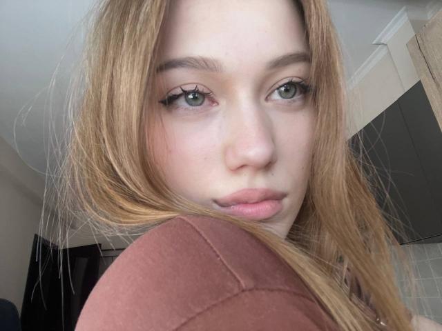 BobbyeWelson's face with long, blonde hair, green eyes, and full pink lips, wearing a brown shirt, looking slightly to the side with a subtle, confident expression.
