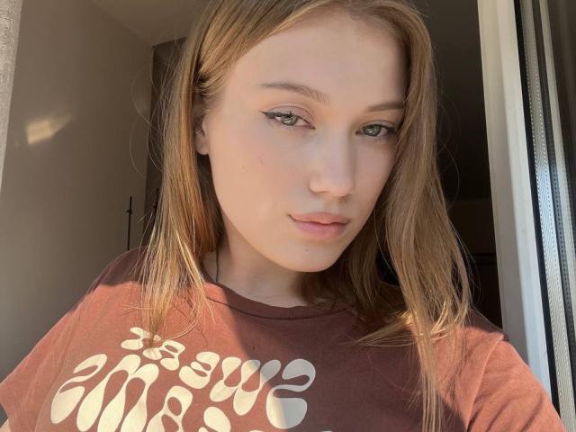 BobbyeWelson with light brown hair, green eyes, and fair skin, wearing a brown shirt, standing in sunlight, showing a subtle smile, and small, slightly parted lips.