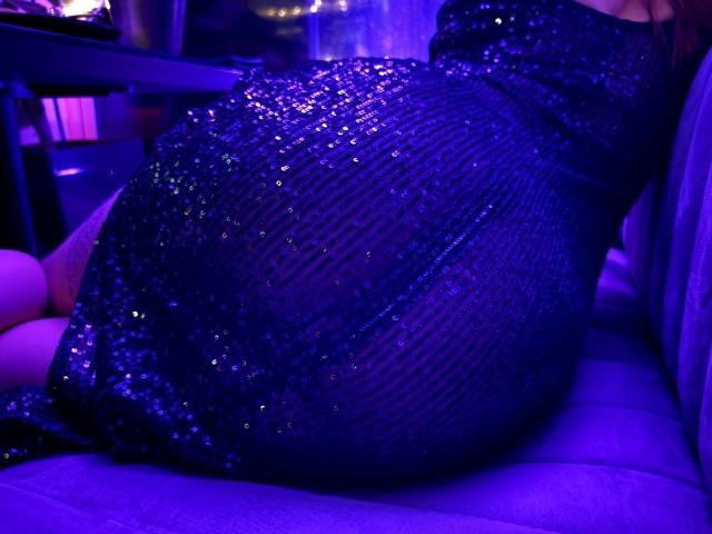 MiraGi est assise sur un canapé éclairé en violet, vêtue d'une robe noire moulante et scintillante avec un motif en mesh. Ses jambes sont visibles sur la gauche..