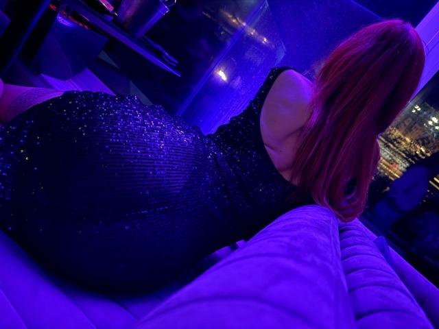MiraGi, con cabello rojo, vestida con un vestido negro brillante, acostada en un sofá morado, mostrando su gran y brillante trasero a la cámara en una habitación iluminada con luz azul..