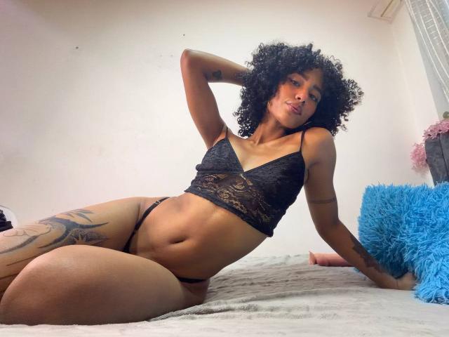 CinthiaWalker with curly black hair, wearing black lace bra and thong, has a tattoo on her left thigh, lying on bed, hand in curly hair, blue pillow to right.