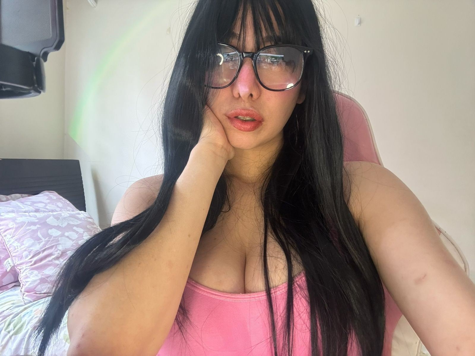 RubyJane mit langen, schwarzen Haaren, Brille, einem pinken Tanktop, das ihre Brüste zeigt, lehnt ihr Kinn in ihre Hand und blickt mit leicht geöffneten, roten Lippen in die Kamera..
