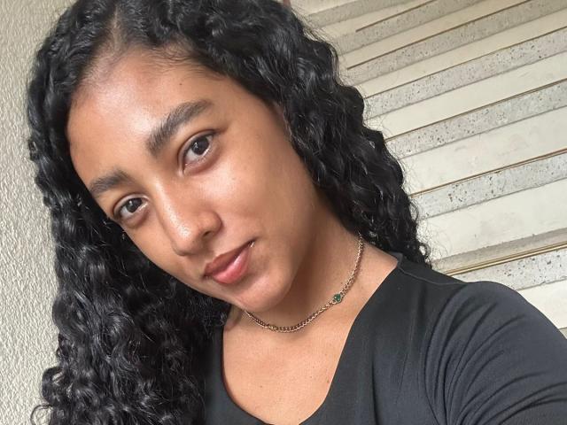 NatalyBlonder has curly black hair, brown eyes, and a cute face with subtle makeup. She's wearing a black top and a thin necklace with a green pendant.