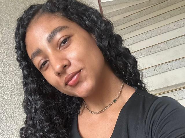NatalyBlonder with curly black hair, medium brown skin, and subtle makeup, wearing a black shirt and silver chain necklace, slightly smiling with soft pink lips.