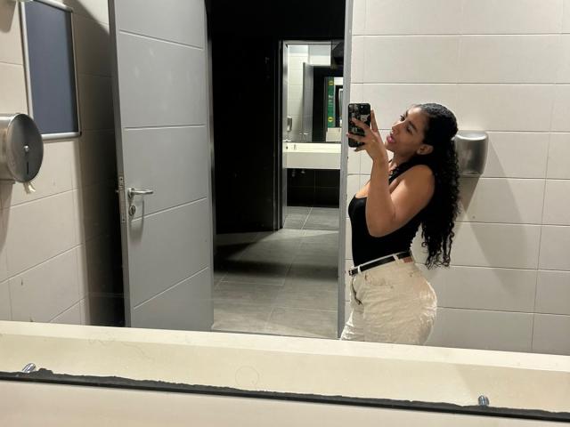 NatalyBlonder with curly black hair, wearing a black tank top and white high-waisted pants, taking a mirror selfie in a bathroom, showing her ass and holding a phone.