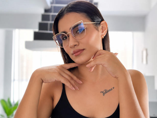 Orianafenix mit gebräunter Haut, schwarzen Haaren und Brille, trägt ein schwarzes Tanktop, zeigt ein Tattoo auf ihrer Brust, ihre Hände sind in der Nähe ihres Halses, und sie hat ein leichtes Lächeln..
