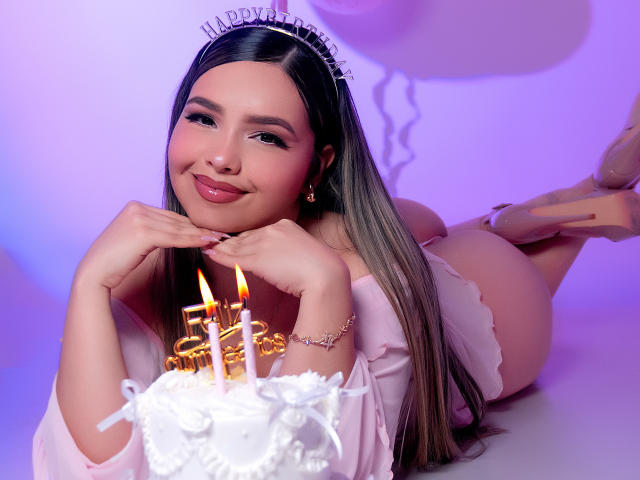 AngelRoussy, lying on her stomach, wearing a pink lace top and white heels, smiling at a cake with lit candles, her hands under her chin, long brown hair, pink makeup.