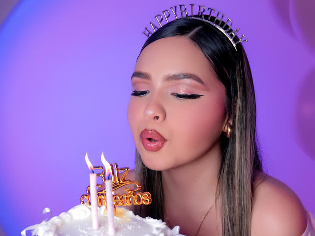 AngelRoussy with pouty lips, wearing a "Happy Birthday" headband, blowing on white frosted cake with four lit candles, her long brown hair, and pink eyeshadow.