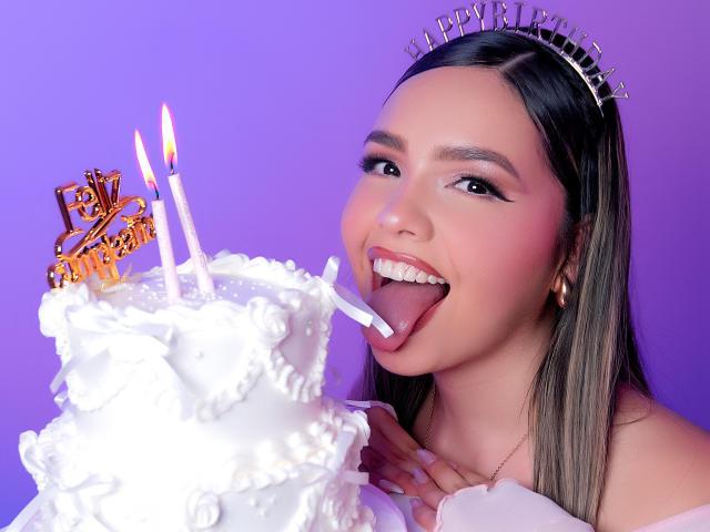 AngelRoussy with long brown hair, wearing a silver tiara, licking a white frosted cake with two lit candles, her tongue out, smiling, against a purple background.