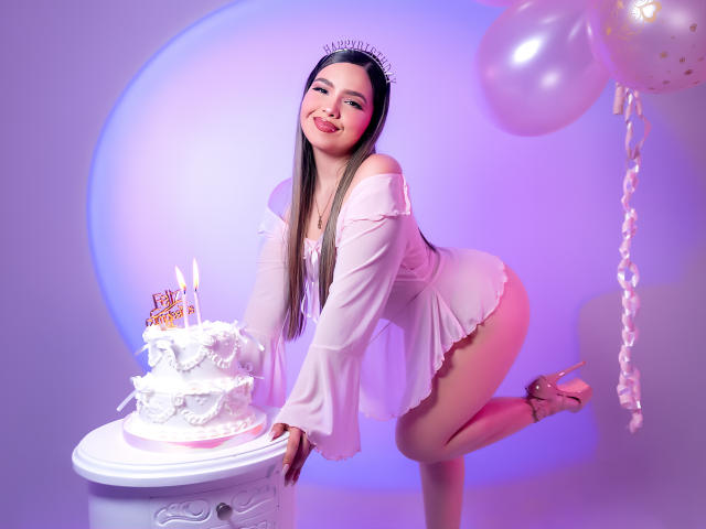 AngelRoussy with long brown hair, wearing a sheer pink dress, bending over a white cake table, pink high heels, and a tiara, smiling at the camera.