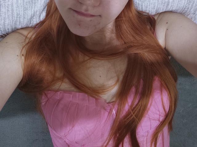 SheritaChieng's red hair, pale skin, and small freckles on her face, wearing a pink top, lying down with arms above her head, visible cleavage, and a subtle smile.