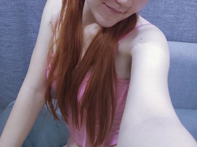 SheritaChieng, avec de longs cheveux roux, de la lingerie rose, une peau pâle, de petites seins, souriante, prenant un selfie, bras droit levé, un coussin bleu-gris derrière elle..