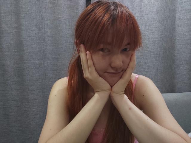 SheritaChieng with red hair, hands on cheeks, smiling slightly, in a pink tank top, sitting with elbows on her knees, looking cute and shy.