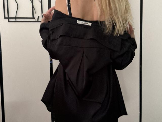 MysteryLaddy with blonde hair, black off-shoulder shirt, revealing black bra straps, holding shirt down, showing back and part of neck, in a slightly provocative, casual pose.