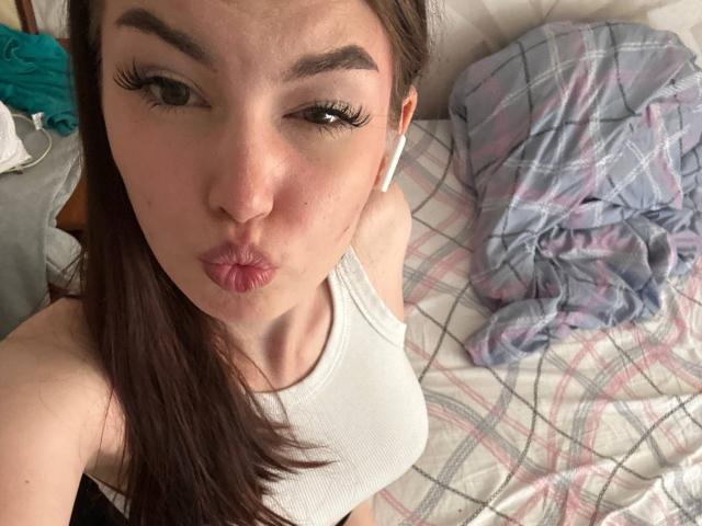 ApoloniaDavick with pouty lips, wearing a white tank top, long brown hair, white earbuds, and thick eyelashes, looking up at the camera. Her tits are small.