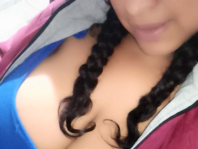 Os seios de ZoehFerraro em uma blusa azul, com tranças caindo, vestindo uma jaqueta rosa e cinza, com decote visível e um leve sorriso. Sua pele é de cor clara..