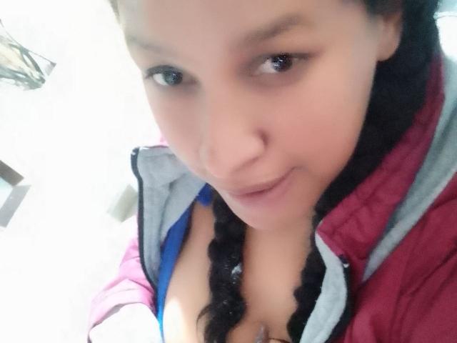 Een selfie van ZoehFerraro waarop haar gezicht en bovenlichaam te zien zijn, met gevlochten zwart haar, een roze en grijze jas, een blauwe bh, en een lichte glimlach, met de nadruk op haar borsten..