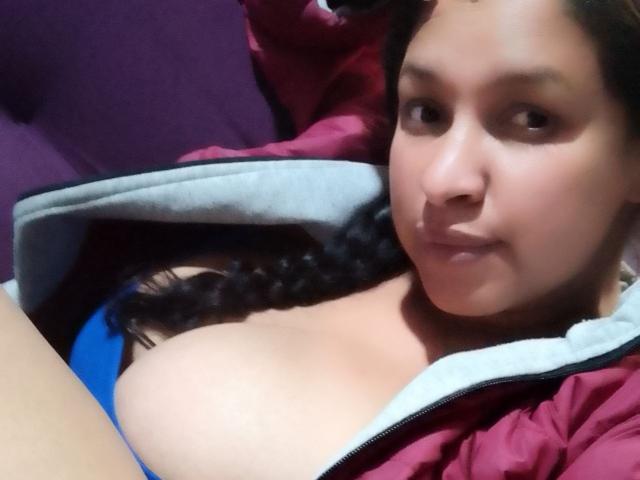 ZoehFerraro has brown eyes and curly black hair, wearing a red and gray jacket, exposing her big tits in a blue top, lying down with a slight smile.